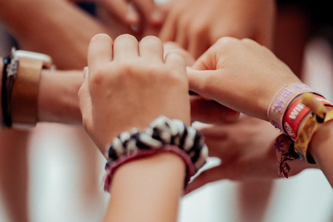 Hände verschiedener Personen legen sich übereinander, viele Armbänder sind sichtbar. Hintergrund unscharf.